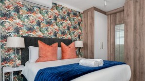 Ein ordentlich gemachtes Bett mit blauer Decke und orangefarbenen Kissen, ein Nachttisch in Weiß mit Lampe sowie eine Wand mit Blumenmuster.