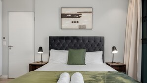 Un lit bien fait, recouvert d’une couverture verte, avec deux serviettes blanches et une tête de lit capitonnée. On y trouve deux lampes de chevet et un tableau encadré au mur.