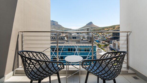 Un balcon avec deux chaises et une petite table, d’où l’on peut admirer un paysage urbain avec des bâtiments et une montagne.