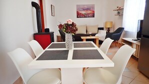 Un salon moderne avec une table à manger blanche, des chaises blanches, un buffet rouge, un canapé noir, un canapé beige, un tableau au mur, une lampe et un téléviseur.