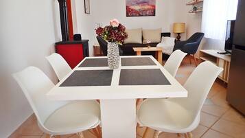 Un salon moderne avec une table à manger blanche, des chaises blanches, un buffet rouge, un canapé noir, un canapé beige, un tableau au mur, une lampe et un téléviseur.
