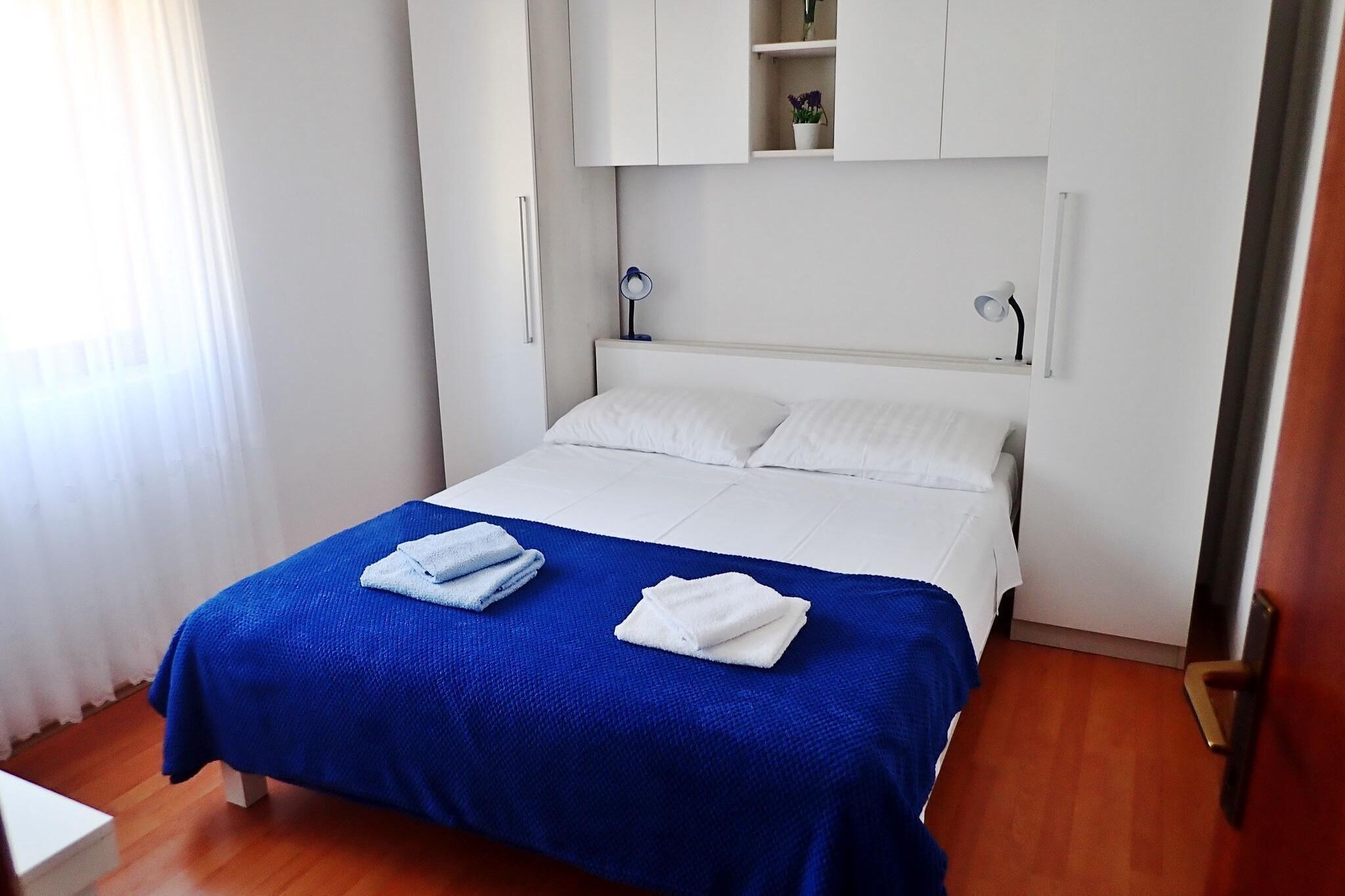 Un lit bien fait, recouvert d’une couvre-lit bleue et blanche, avec des oreillers blancs et deux serviettes pliées. Au-dessus du lit, on trouve une armoire blanche avec une petite étagère sur laquelle sont posés un vase et une lampe.