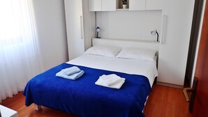 Un lit bien fait, recouvert d’une couvre-lit bleue et blanche, avec des oreillers blancs et deux serviettes pliées. Au-dessus du lit, on trouve une armoire blanche avec une petite étagère sur laquelle sont posés un vase et une lampe.