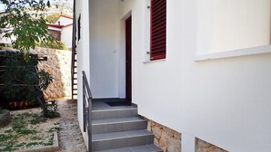 Un escalier gris mène à une porte ornée d’un volet rouge, adossée à un mur de pierre, et devant se trouve un petit jardin avec des plantes et un arbre.
