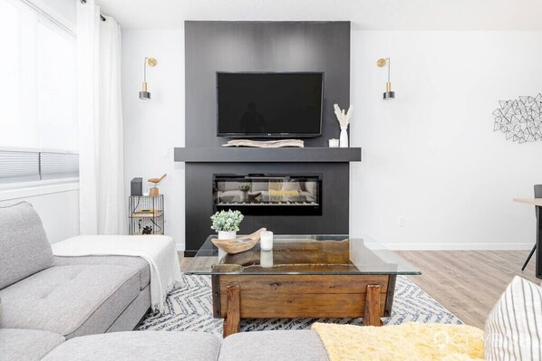 Un soggiorno moderno con una TV a schermo piatto montata sopra un caminetto, un tavolino da caffè in vetro e un divano grigio.