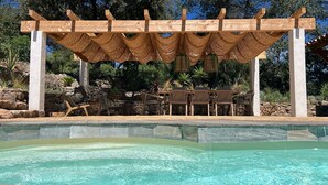 Un espace aménagé au bord de la piscine, doté d’une pergola, de sièges d’extérieur et d’un mur en pierre en arrière-plan.