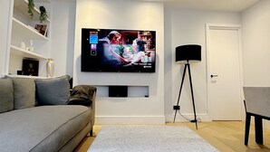A modern living room with a grey sofa, a patterned ottoman, a flat-screen TV mounted on the wall, a standing lamp, and a small table with a chair. - Londwell - Chelsea Garden Flat - Private Oasis (London)