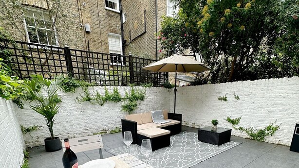 An outdoor patio with a table set for dining, surrounded by chairs and a cushioned bench. - Londwell - Chelsea Garden Flat - Private Oasis (London)