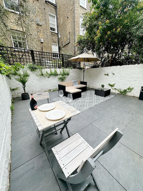Relax in a serene outdoor space with a long table, cushioned chairs, and a bench, shaded by a large umbrella.