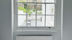 A room with a large window, a radiator below, and a view of a garden and buildings outside. - Londwell - Chelsea Garden Flat - Private Oasis (London)