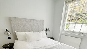 A neatly made bed with a grey and white blanket, two bedside tables with lamps, a window with a view of greenery, and a radiator under the window. - Londwell - Chelsea Garden Flat - Private Oasis (London)