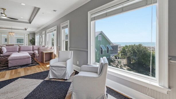 A spacious living room with a large window, a sectional sofa, and a view of a green house and beach. - 4 Bed 5 Bath Luxury Beach Front Home w/Ocean Views (Tybee Island)