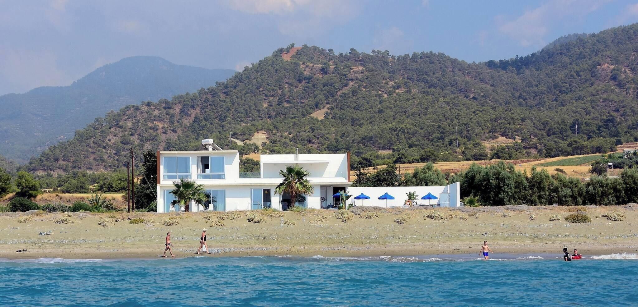 A coastal building with a modern design, a beach in the foreground, and a mountainous backdrop.