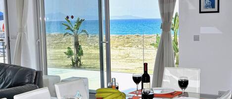 A room with a dining table set for two, a bottle of wine, and a glass of red wine. There is a view of the beach and the sea through large windows.