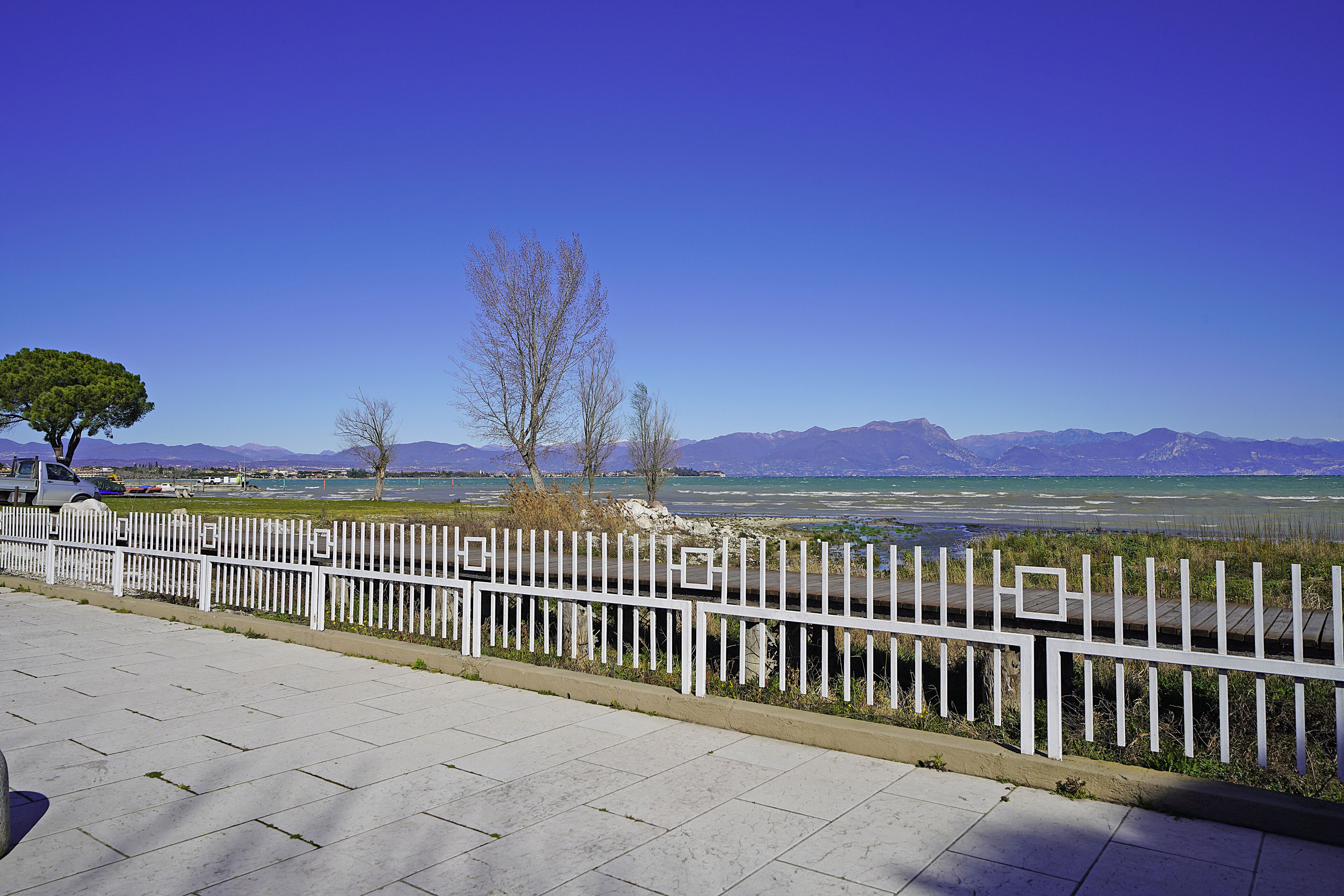 A paved walkway with a white fence, a tree, and a view of mountains and water.