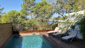 Ein Schwimmbecken mit Holzterrasse und Liegestühlen, umgeben von Bäumen und einer weißen Mauer.