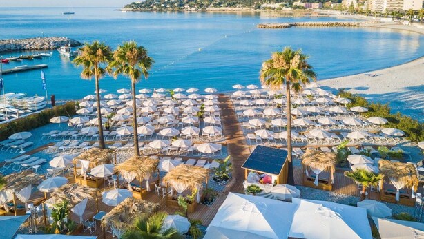 Ein Strandhotel mit vielen Liegestühlen und Sonnenschirmen, einem Poolbereich und Blick auf das Meer und Gebäude in der Ferne.