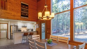 A wooden interior with a dining table, chairs, a chandelier, and a view through large windows. - RR070 | Updated open concept home close to ranch amenities and hiking trails. (Black Butte Ranch)