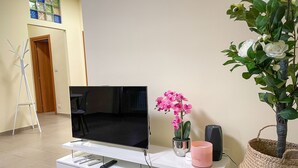 Ett vardagsrum med en platt-TV på ett vitt TV-bord, en rosa orkidé, en vit vas och en flätad korg.