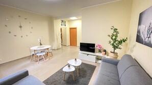 Ett vardagsrum med en grå soffa, ett litet runt bord med en växt, en TV-bänk med en platt-TV och en matplats med ett bord och stolar.