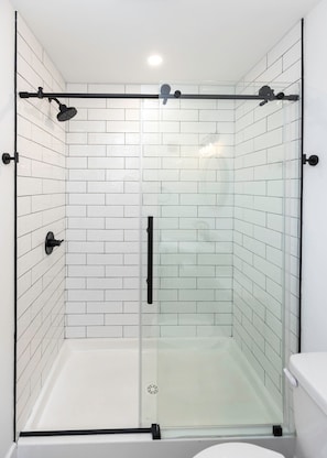 A glass-enclosed shower stall with white tiled walls and a white toilet visible to the right.