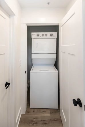 A laundry room with a stacked washer and dryer, white doors, and a wooden floor. - Jeffreys Bay - Loft #3 - 8 min walk to beach (Virginia Beach)