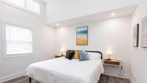 A bedroom with a bed, two bedside tables, a wall-mounted picture, and a window with blinds. - Jeffreys Bay - Loft #3 - 8 min walk to beach (Virginia Beach)