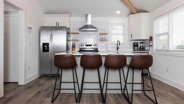 A modern kitchen with white cabinetry, a stainless steel refrigerator, a gas range, and four bar stools. - Malibu - Loft #4 - 8 min walk to the beach (Virginia Beach)