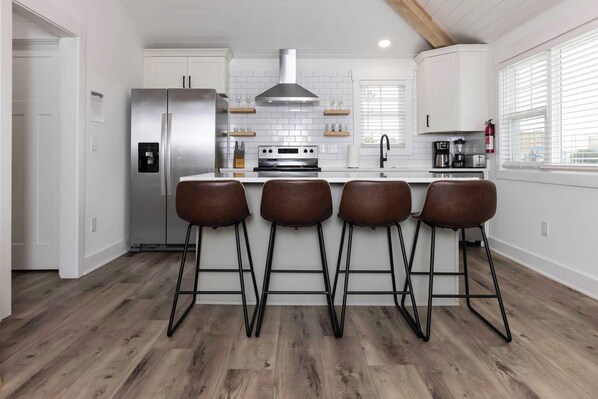 A modern kitchen with white cabinetry, a stainless steel refrigerator, a gas range, and four bar stools.