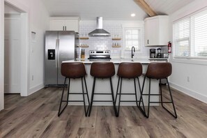 Una cocina moderna con armarios blancos, un refrigerador de acero inoxidable, una cocina a gas y cuatro taburetes de bar.