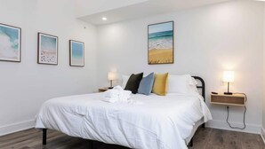 A hotel room with a bed, two bedside tables, a nightstand, a wall-mounted lamp, and three framed pictures. - Malibu - Loft #4 - 8 min walk to the beach (Virginia Beach)