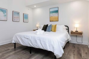 A hotel room with a bed, two bedside tables, a nightstand, a wall-mounted lamp, and three framed pictures. - Malibu - Loft #4 - 8 min walk to the beach (Virginia Beach)