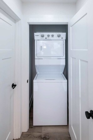 A compact laundry room with a stacked washer and dryer, white walls, and a door with a black handle. - Malibu - Loft #4 - 8 min walk to the beach (Virginia Beach)