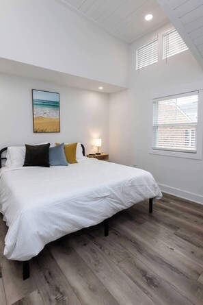 A bedroom with a bed, a nightstand, a lamp, a painting of a beach, and a window with blinds.