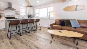 A modern kitchen with white cabinets, a stove, and a bar area with stools. There's a brown leather sofa and a round table with a remote control. - Malibu - Loft #4 - 8 min walk to the beach (Virginia Beach)