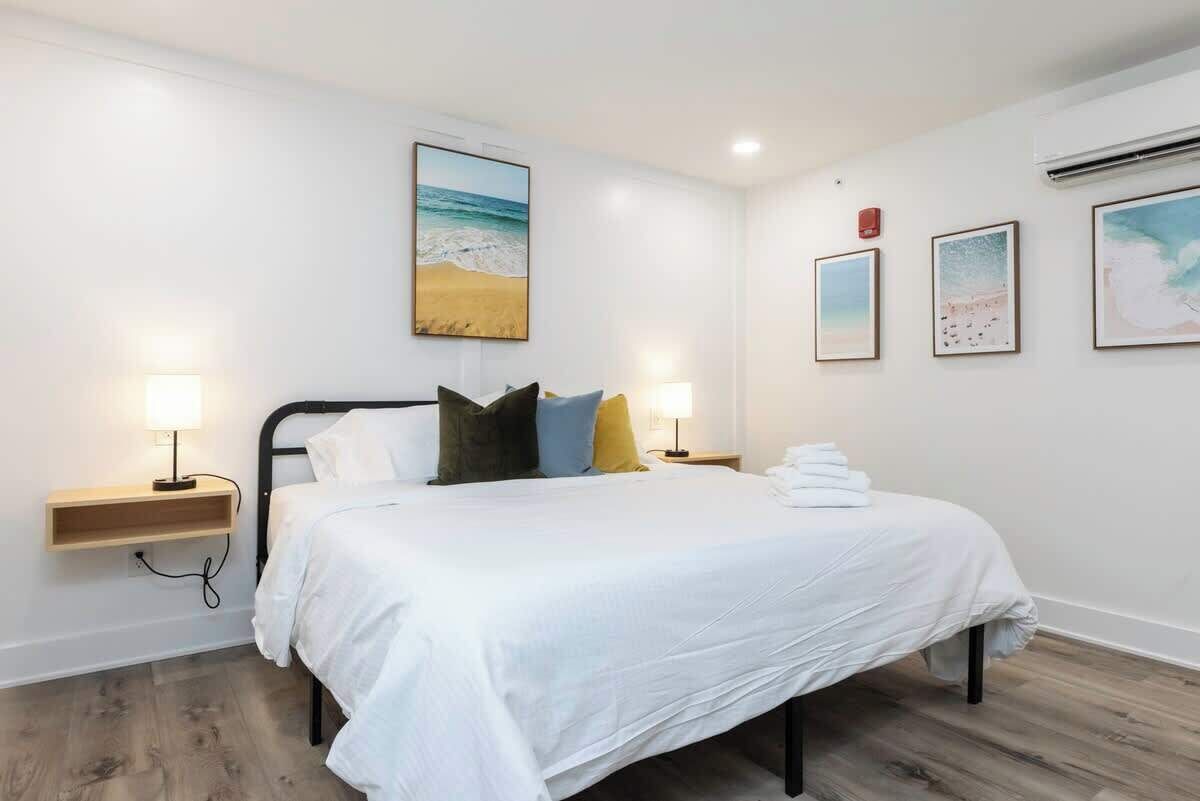 Une chambre à coucher avec un lit, des lampes de chevet, une étagère fixée au mur et un tableau sur le thème de la plage.