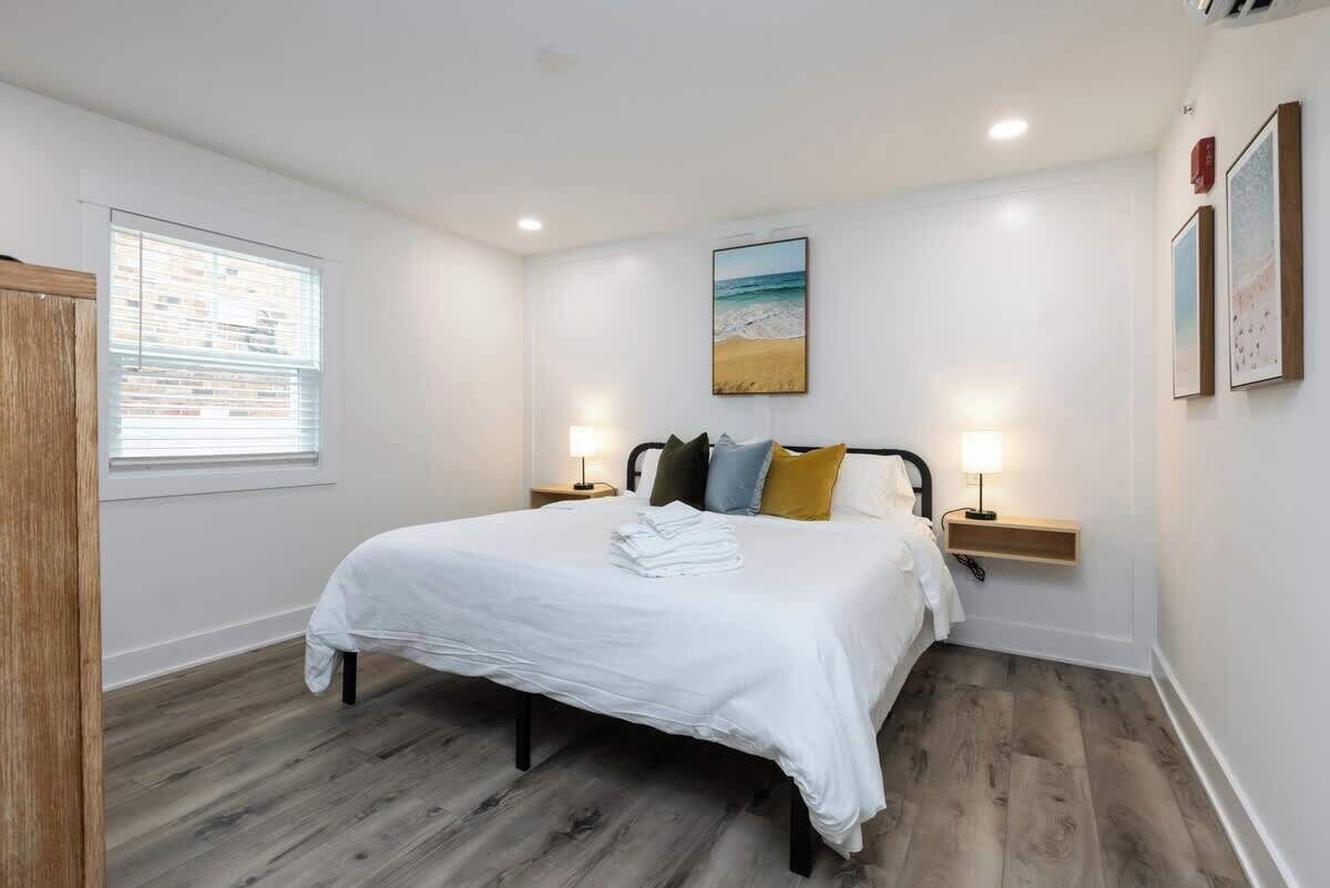 Une chambre à coucher avec un lit, deux tables de chevet, une fenêtre avec des stores, un tableau au mur et un plancher en bois.