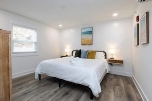 Quarto com cama, duas mesas de cabeceira, janela com persianas, um quadro na parede e piso de madeira.