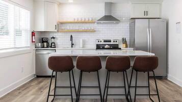 Une cuisine moderne avec des meubles blancs, un réfrigérateur en acier inoxydable, un îlot central doté de tabourets et une fenêtre avec des stores.