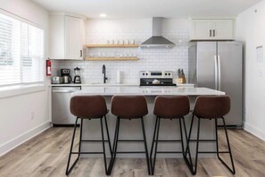 Eine moderne Küche mit weißen Schränken, einem Edelstahl-Kühlschrank, einer Kochinsel mit Hockern und einem Fenster mit Jalousien.
