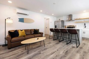 A modern living room with a brown leather sofa, a round coffee table, and a kitchen area with a white refrigerator, stove, and bar stools. - Rincon - Loft #6 - 8 min walk to the beach (Virginia Beach)