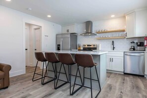 A modern kitchen with a central island, bar stools, stainless steel appliances, and white cabinetry. - Rincon - Loft #6 - 8 min walk to the beach (Virginia Beach)