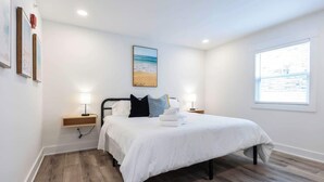 Een slaapkamer met een bed, bedlampen, een nachtkastje, een raam met uitzicht en een lijst met een afbeelding aan de muur.