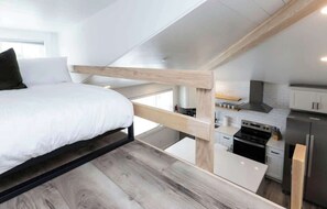 A modern loft bedroom with a bed, wooden beam ceiling, and a kitchen area below.