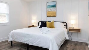 Una cama bien hecha con ropa blanca, dos lámparas de noche, un cuadro en la pared de una playa y suelo de madera.