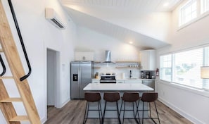 Eine moderne Küche mit weißen Schränken, einem Edelstahl-Kühlschrank, einer zentralen Kochinsel mit Barhocker und einem großen Fenster, das für natürliches Licht sorgt.