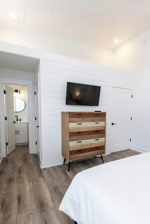 Ein Schlafzimmer mit einem Bett, einer Holzkombination, einem an der Wand montierten Fernseher und einem durchgehenden Türrahmen einsehbaren Badezimmer.
