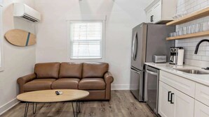 A compact living space with a kitchenette, a brown leather sofa, a small round table, and a window with blinds. - Pipeline - Loft #9 - 8 min walk to the beach (Virginia Beach)