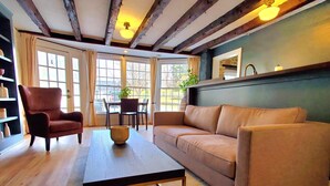 Un salón acogedor con suelo de madera, un sofá de cuero marrón, una mesa de centro oscura y un sillón rojo. Hay una ventana con cortinas, una planta en maceta y un cuadro en la pared.