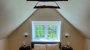 Un dormitorio con techo inclinado, un ventanal grande, dos mesitas de noche con lámparas, una cama con cubrecama color beige y marrón, y suelo de madera.